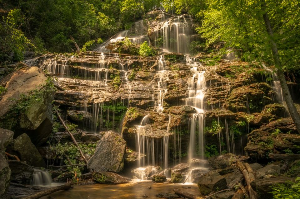 Water fans across a wide tiered rock face in long-exposure silk threads, surrounded by sunlit green forest, pooling among mossy boulders below.