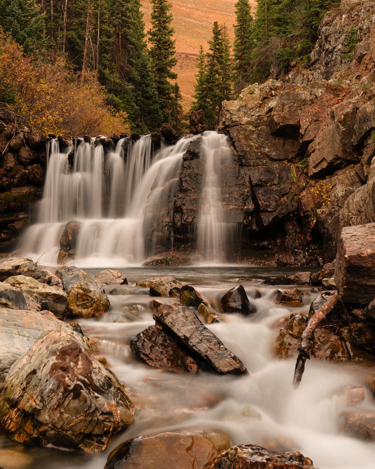 Long-exposure waterfall splits over dark rock ledges into a boulder-strewn pool, flanked by autumn conifers and orange shrubs on a mountain slope.