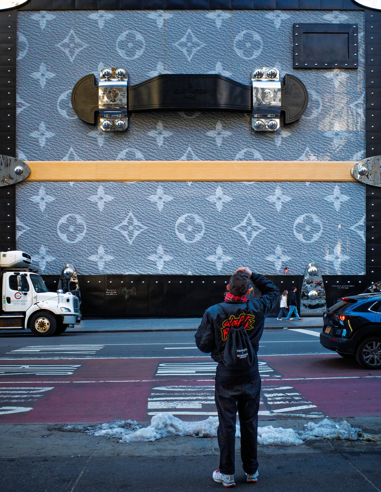 Person in a black jacket stands at a crosswalk facing a building wrapped as a giant Louis Vuitton trunk, slush on the curb.