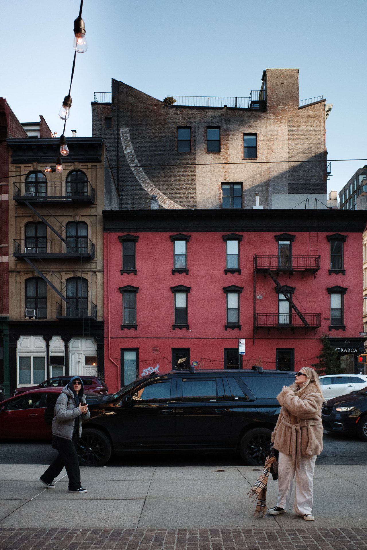 Two pedestrians on a sidewalk in front of a red brick building with fire escapes; one looks up, string lights hang overhead.
