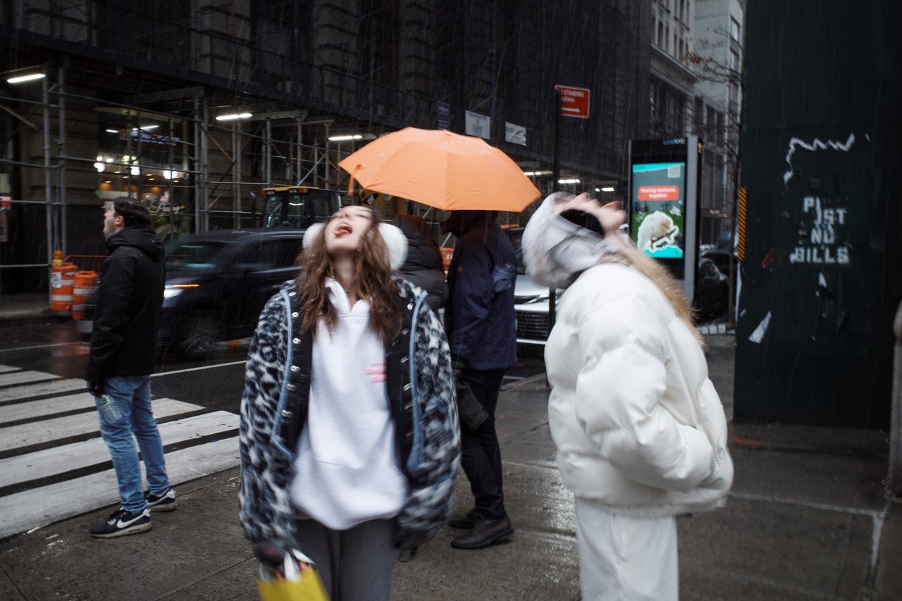 Two women on a rainy city sidewalk look upward, one in a leopard-print coat laughing, as a person holds an orange umbrella behind them.