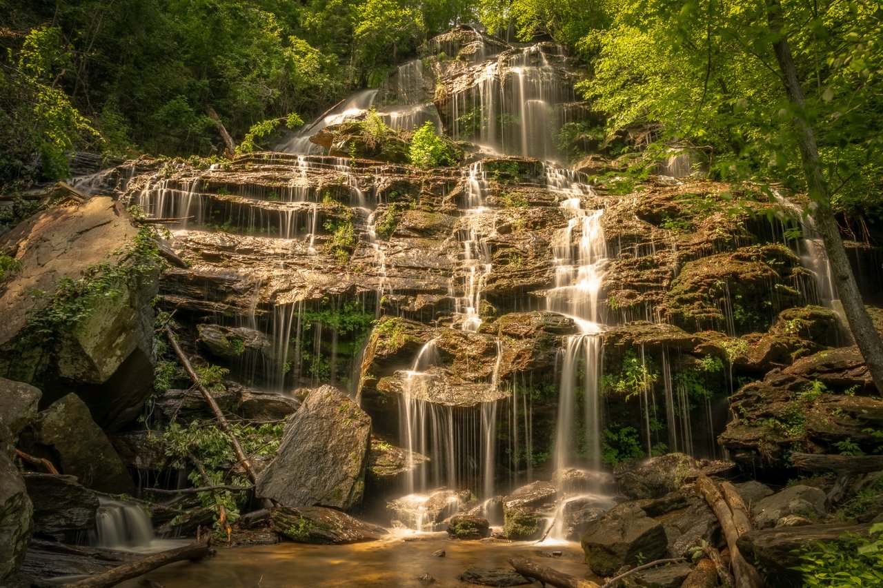 Water fans across a wide tiered rock face in long-exposure silk threads, surrounded by sunlit green forest, pooling among mossy boulders below.