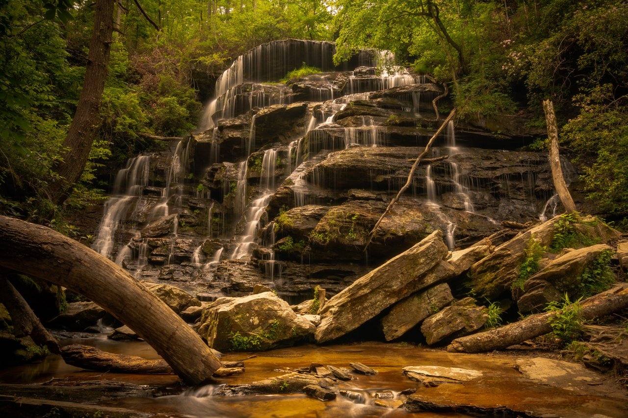 Long-exposure waterfall cascades down wide layered rock shelves into a tannin-stained pool, fallen logs and boulders filling the foreground.