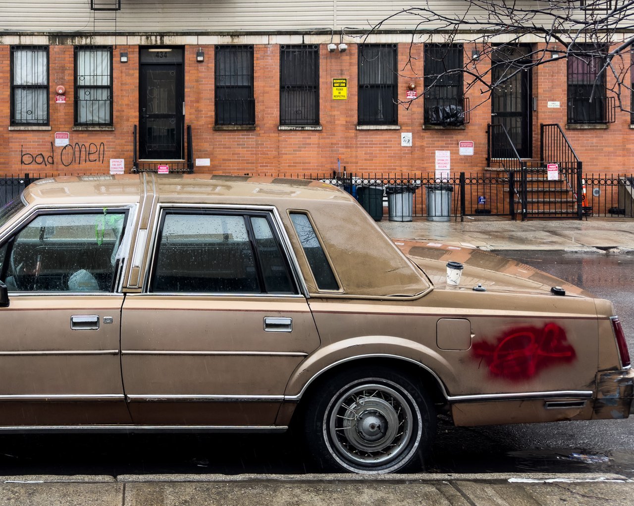 Gold 1980s sedan parked on a wet city street, red graffiti on its trunk, coffee cup on roof, brick rowhouses behind with "bad omen" on the wall.