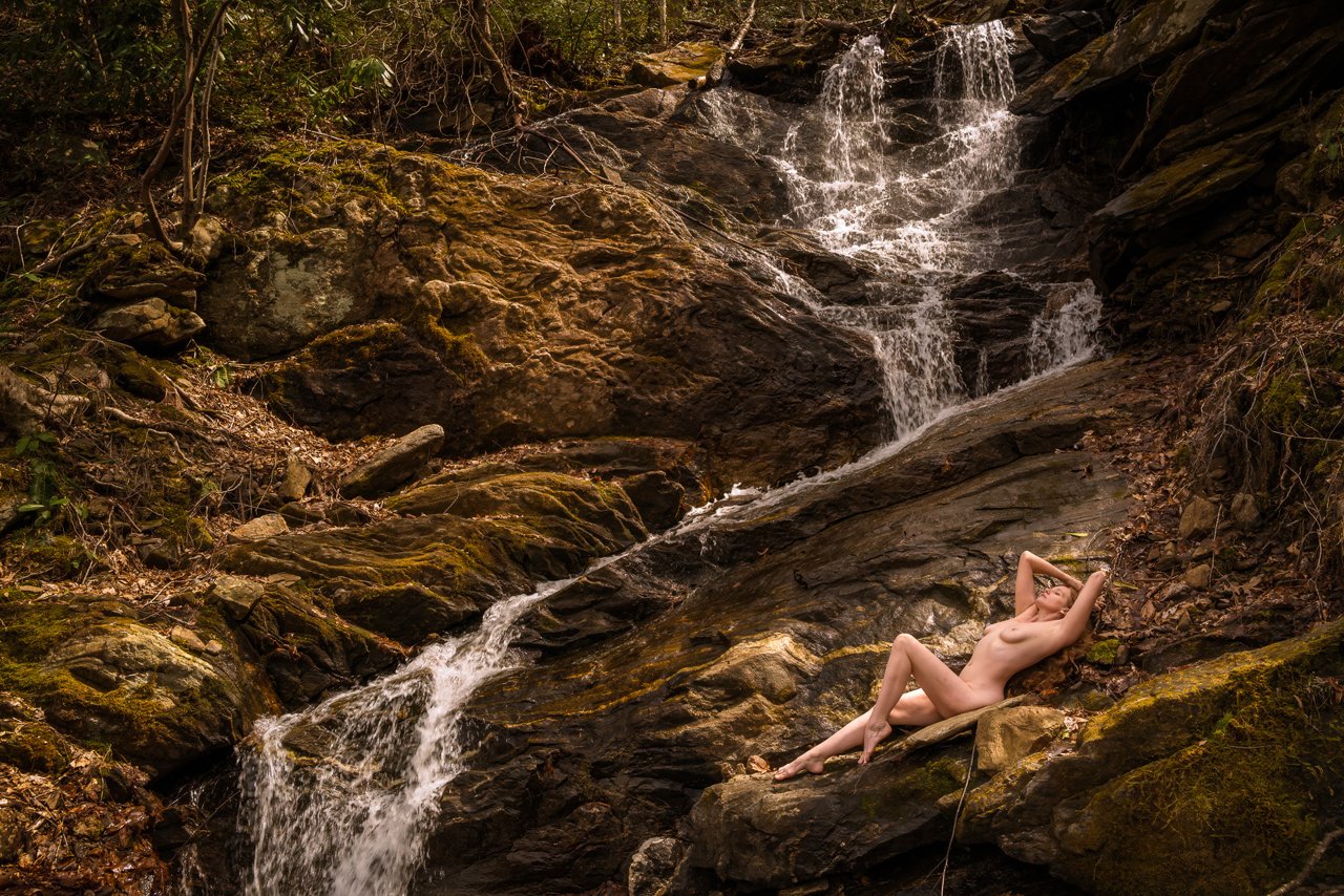 Nude woman reclines on a moss-edged boulder beside a cascading rocky waterfall, arms raised behind her head, forest foliage in background.