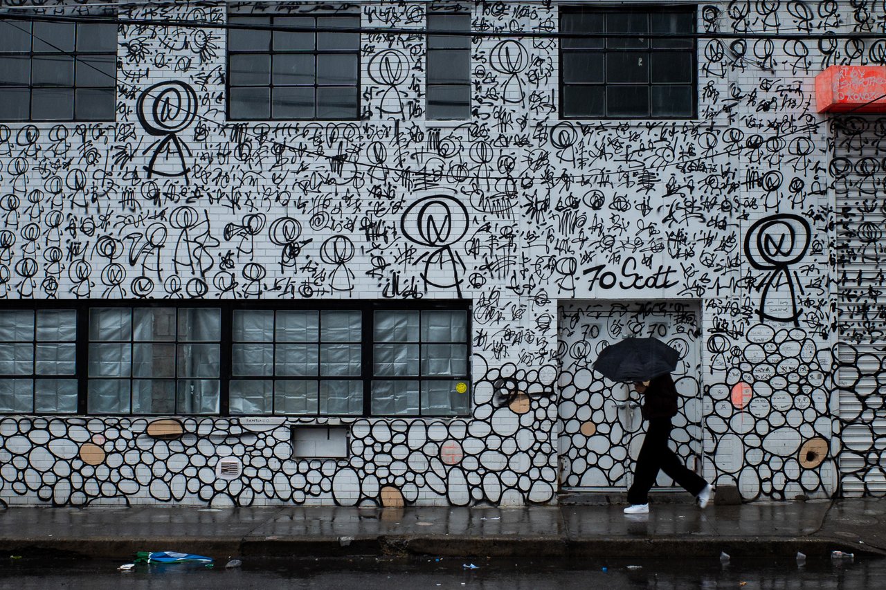 Person with a black umbrella walks past a white brick building covered in black stick-figure and abstract graffiti murals on a rainy day.