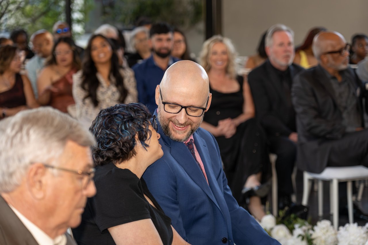 Bald man in a navy suit smiles down at a woman whispering in his ear, seated among rows of formally dressed guests at an indoor ceremony.