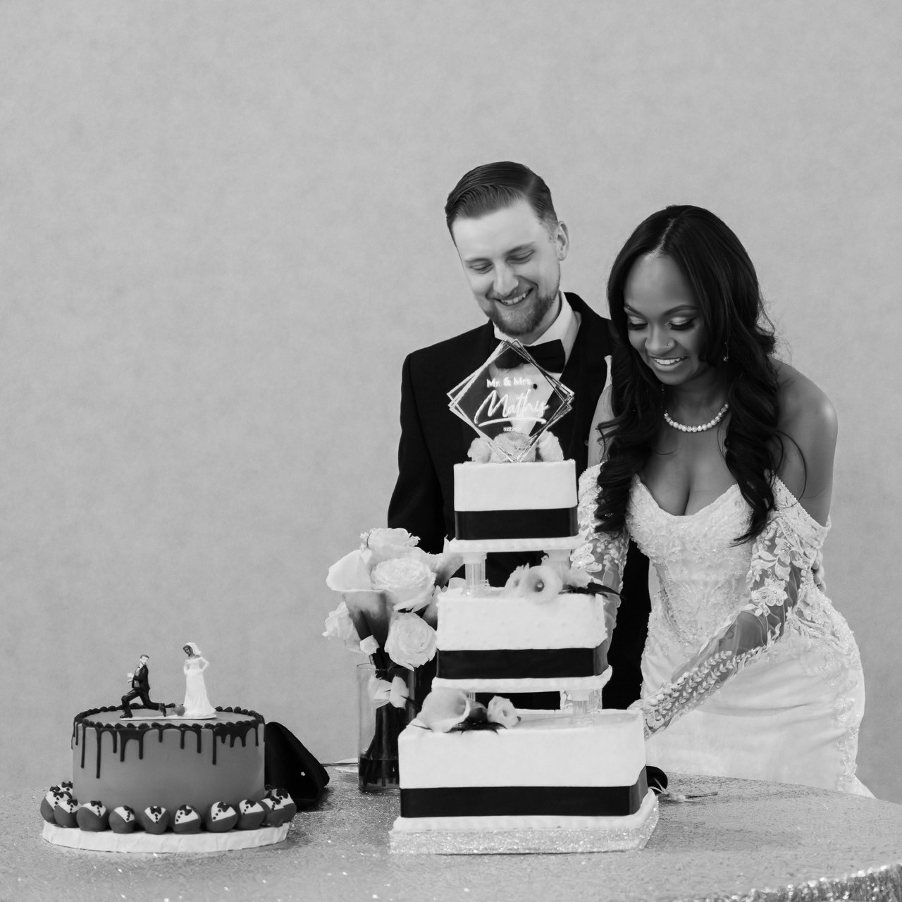 Bride in lace gown and groom in tuxedo cut a tiered wedding cake beside a smaller drip cake with figurine topper, black and white.