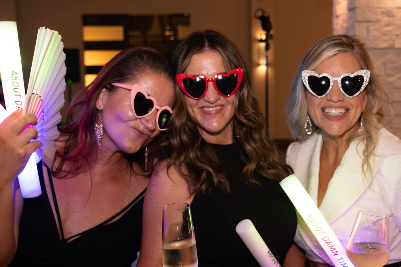 Three women wearing oversized heart-shaped sunglasses pose indoors at a party, holding glow sticks, a paper fan, and champagne flutes under warm event lighting.