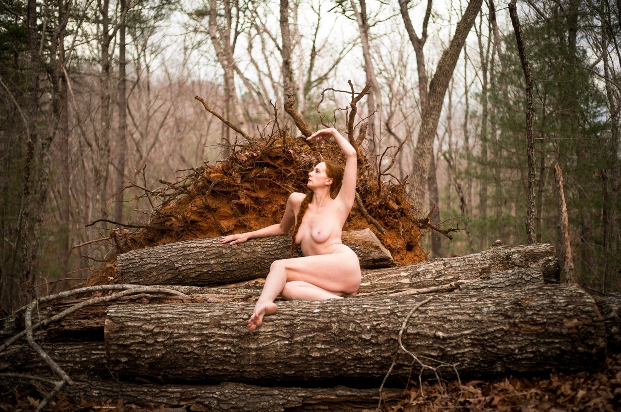Nude woman seated on a fallen log, one arm raised, beside an uprooted tree's exposed root mass in a bare winter forest.